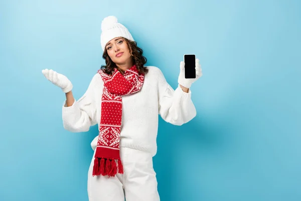 Confused young woman in winter outfit holding mobile phone with blank screen on blue — Stock Photo