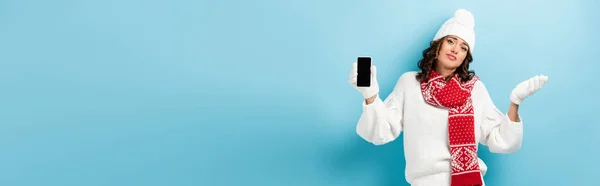 Панорамна концепція заплутаної молодої жінки в зимовому вбранні, що тримає мобільний телефон з порожнім екраном на синьому — стокове фото