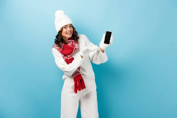 Молода жінка в зимовому вбранні вказує рукою на мобільний телефон з порожнім екраном на синьому — стокове фото