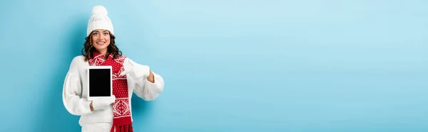 Eine fröhliche Frau im Winteroutfit zeigt mit dem Finger auf ein digitales Tablet mit leerem Bildschirm auf blauem Hintergrund — Stockfoto