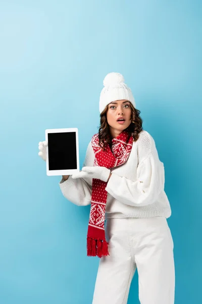 Surprised young woman in winter outfit holding digital tablet with blank screen on blue — Stock Photo