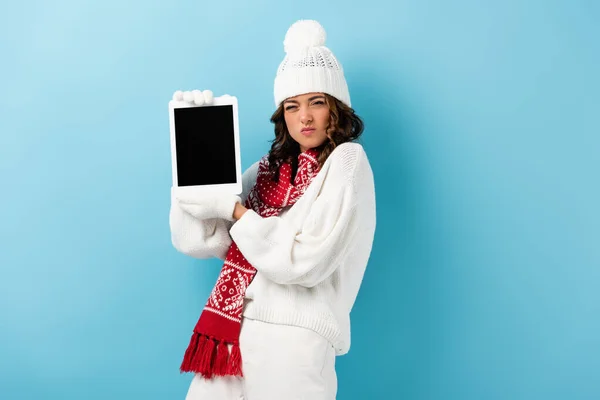 Angry young woman in winter outfit holding digital tablet with blank screen on blue — Stock Photo