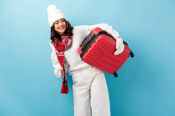 Fröhliche junge Frau im Winteroutfit mit rotem Koffer auf blauem — Stockfoto