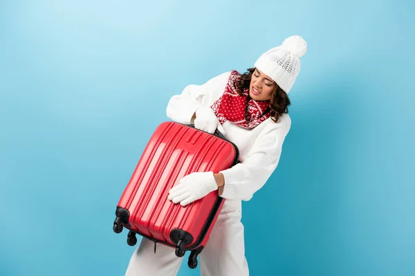 Mujer joven enojada en traje de invierno llevando maleta roja en azul - foto de stock