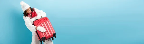Panoramic shot of angry young woman in winter outfit carrying red suitcase on blue — Stock Photo