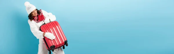 Tiro panorámico de mujer joven enojada en traje de invierno llevando maleta roja en azul - foto de stock