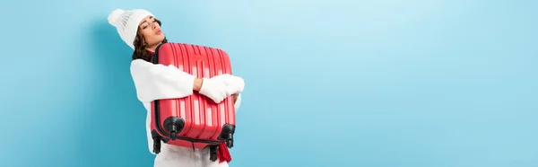 Panoramic shot of young woman in winter outfit carrying heavy suitcase on blue — Stock Photo