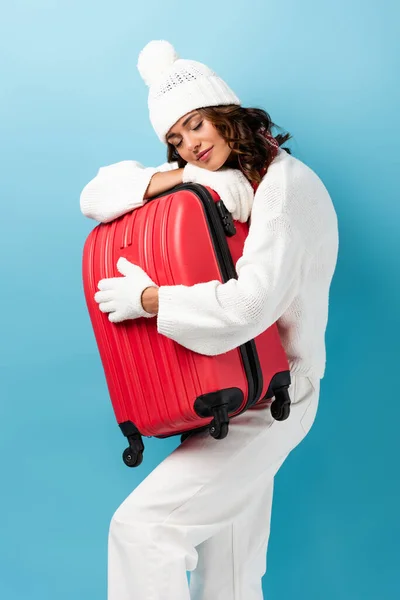 Young woman with closed eyes, in winter outfit carrying suitcase on blue — Stock Photo