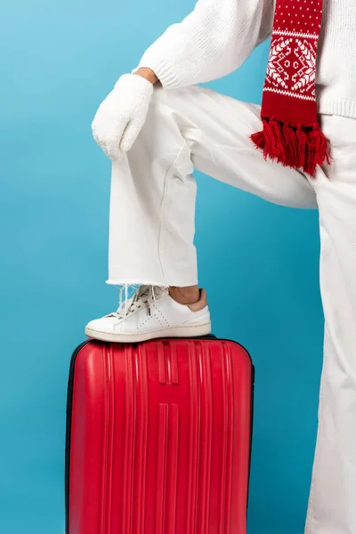 Cropped view of young woman in winter outfit standing on suitcase isolated on blue — Stock Photo