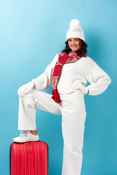 Joyful young woman in winter outfit standing on suitcase on blue — Stock Photo