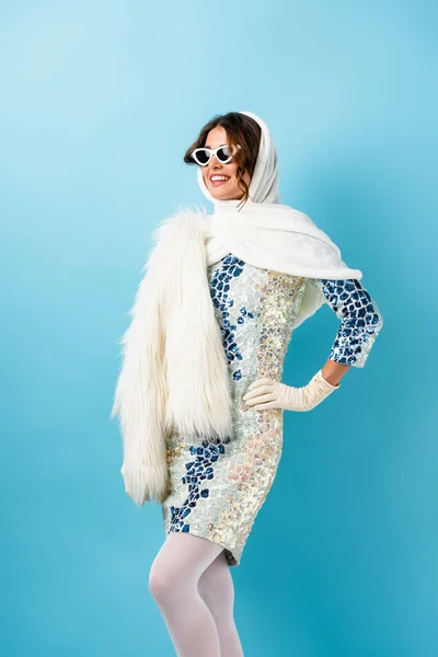 Mujer alegre en gafas de sol y vestido de pie con la mano en la cadera en azul - foto de stock