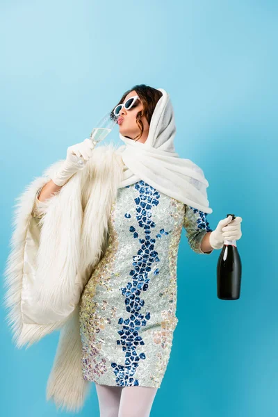 Mulher elegante em óculos de sol segurando garrafa e bebendo champanhe em azul — Fotografia de Stock