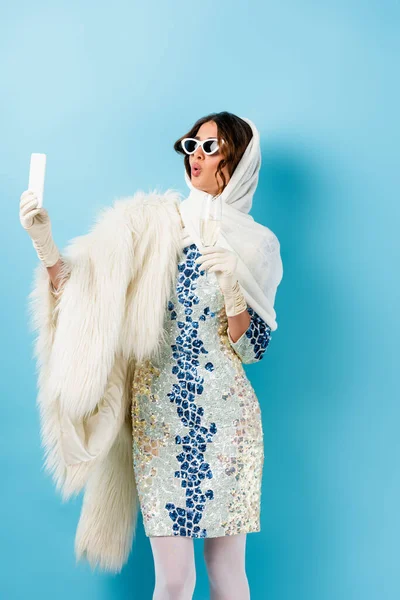 Стильна жінка в сонцезахисних окулярах тримає келих шампанського і приймає селфі на синьому — стокове фото