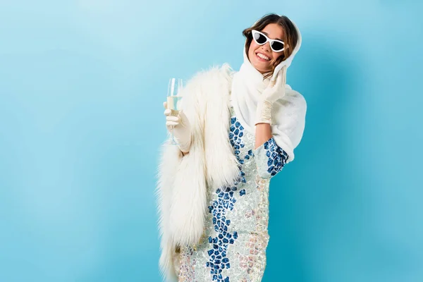 Радісна жінка в сонцезахисних окулярах тримає келих шампанського і розмовляє на смартфоні на синьому — стокове фото