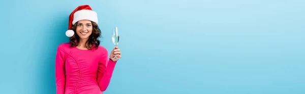 Horizontal crop of young happy woman in santa hat holding glass of champagne on blue — Stock Photo