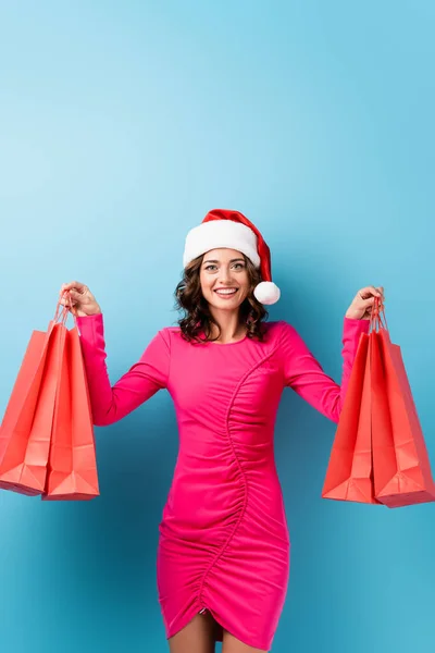 Mulher alegre em vestido rosa e chapéu de santa segurando sacos de compras em azul — Fotografia de Stock