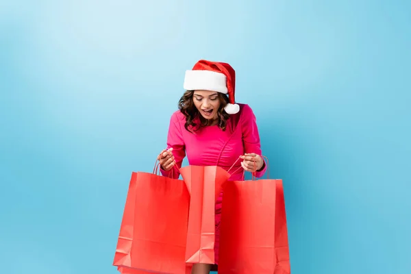 Excited young brunette woman in santa hat looking at shopping bags on blue — Stock Photo