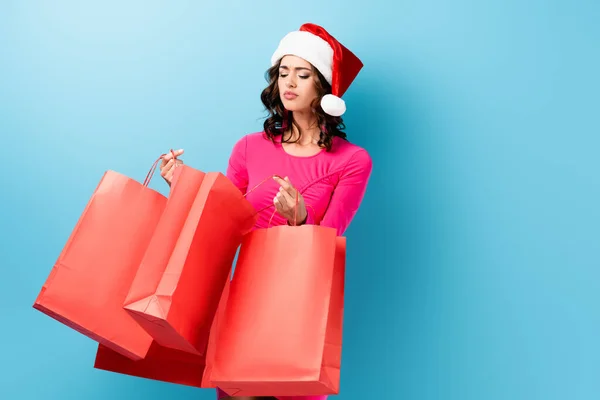 Joven morena mujer en santa hat mirando las bolsas de compras en azul - foto de stock