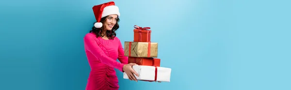 Horizontales Bild einer jungen fröhlichen Frau mit Weihnachtsmütze und Kleid, die umwickelte Geschenke auf blauem Grund hält — Stockfoto