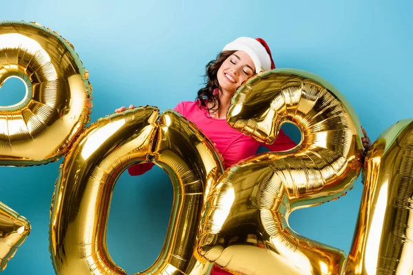 Mulher alegre em santa chapéu perto de balões com 2021 números em azul, conceito de ano novo — Fotografia de Stock