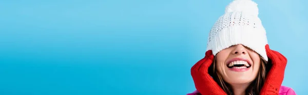 Panoramic shot of joyful woman in red gloves covering eyes while touching white hat isolated on blue — Stock Photo