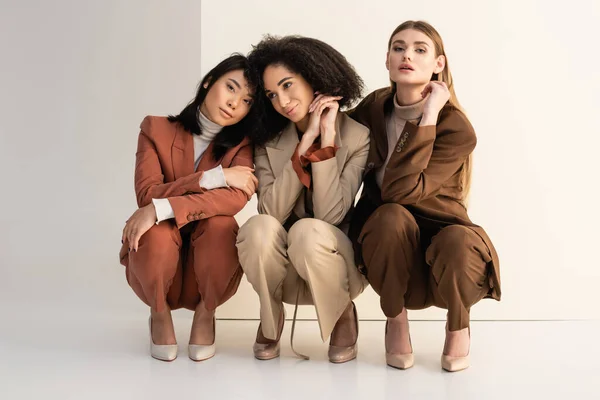 Young interracial women in trendy suits sitting and posing on white — Stock Photo