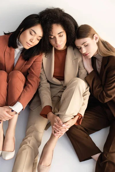 Vista de ángulo alto de las mujeres multiculturales con los ojos cerrados en trajes de moda mirando a la cámara en blanco - foto de stock