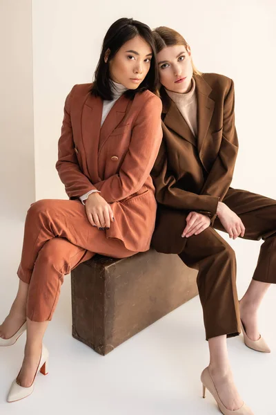 Interracial women in trendy suits sitting on suitcase on white — Stock Photo