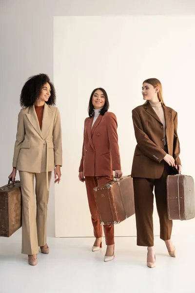 Full length of interracial women in trendy suits holding suitcases on white — Stock Photo