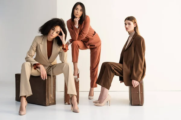 Longitud completa de las mujeres multiculturales con estilo en trajes sentados en maletas en blanco - foto de stock