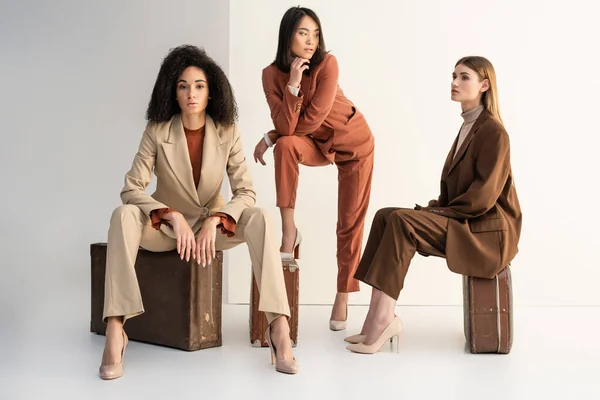 Full length of multicultural women in trendy suits sitting on luggage on white — Stock Photo