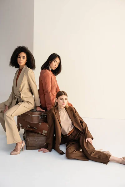 Full length of young multicultural women in trendy suits sitting on suitcases on white — Stock Photo