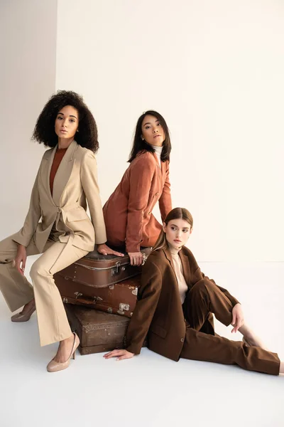 Full length of multicultural women in trendy suits sitting on suitcases on white — Stock Photo