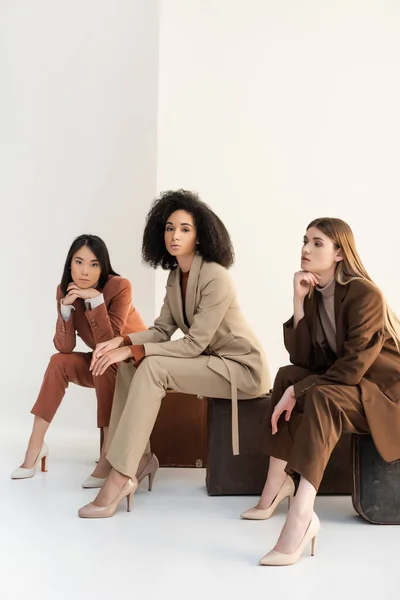 Longitud completa de las mujeres multiculturales en ropa formal de moda sentado en maletas en blanco - foto de stock