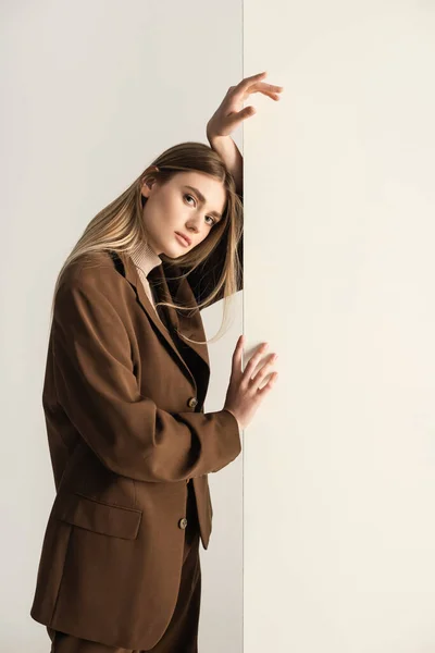 Jovem mulher loira em elegante terno marrom olhando para a câmera enquanto se inclina na parede no branco — Fotografia de Stock