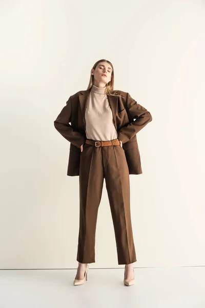 Longitud completa de la joven rubia en elegante traje marrón de pie con las manos en las caderas en blanco - foto de stock
