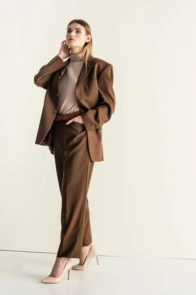Блондинка в стильном коричневом костюме в полный рост смотрит в камеру, стоя с рукой в кармане на белом — стоковое фото