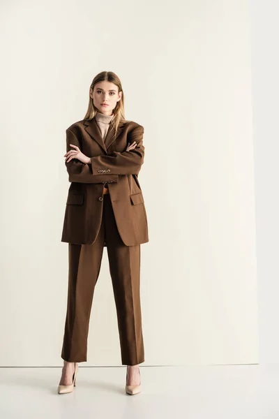 Longitud completa de la joven mujer rubia en elegante traje marrón mirando a la cámara mientras está de pie con los brazos cruzados en blanco - foto de stock