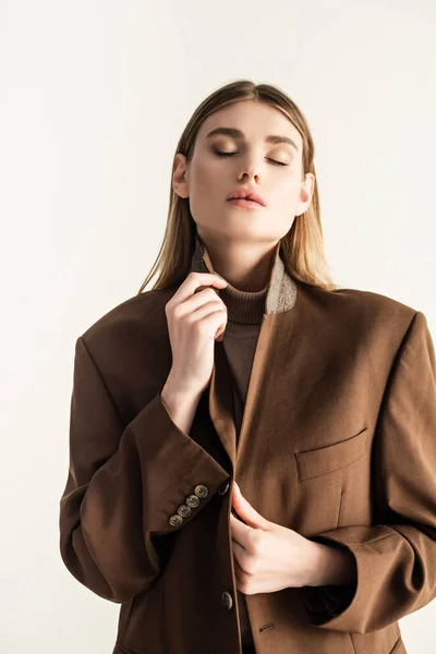 Mujer rubia joven con los ojos cerrados ajustando elegante chaqueta marrón aislado en blanco — Stock Photo