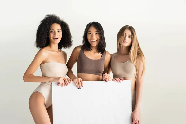 Alegre interracial las mujeres en beige ropa interior celebración en blanco cartel aislado en blanco - foto de stock