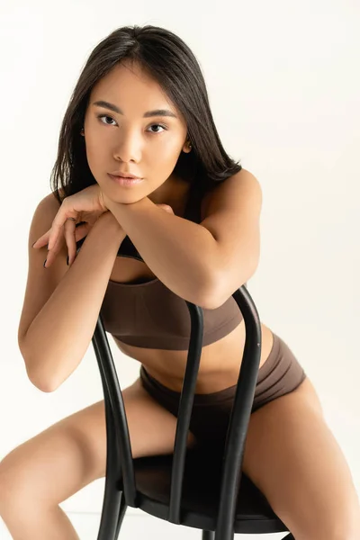 Brünette asiatische Frau in beiger Unterwäsche schaut in die Kamera, während sie auf einem Stuhl isoliert auf weiß posiert — Stockfoto