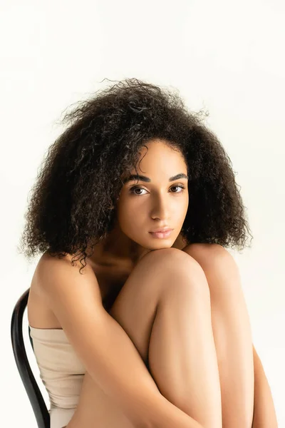 Frisé afro-américain femme regardant caméra tout en posant sur chaise isolée sur blanc — Photo de stock