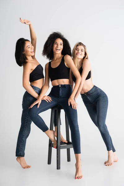 Glückliche interrassische Frauen in Jeans posieren in der Nähe von Hockern auf weißem Grund — Stockfoto