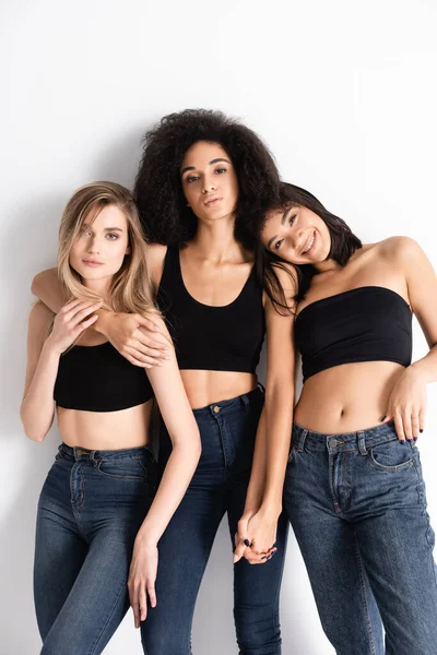 Young interracial women in denim jeans posing with cheerful asian model on white — Stock Photo