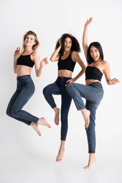 Excité interracial les femmes en denim jeans sautant et posant sur blanc — Photo de stock