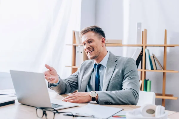 Hombre de negocios sonriente que tiene chat de vídeo en el portátil cerca de los auriculares y papeles en primer plano borrosa - foto de stock