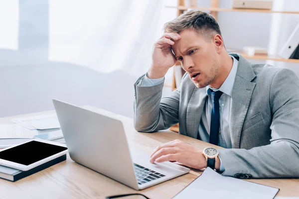 Nachdenklicher Geschäftsmann mit Laptop in der Nähe von Zeitungen und digitalem Tablet im unscharfen Vordergrund — Stockfoto