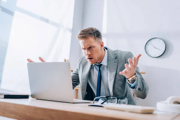 Confused businessman looking at laptop near eyeglasses and headphones on burred foreground — Stock Photo