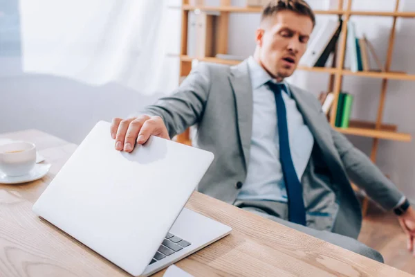 Бизнесмен закрывает ноутбук возле чашки кофе на размытом фоне в офисе — стоковое фото