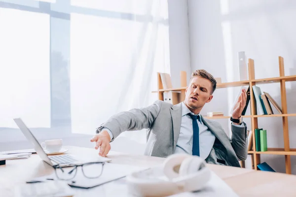 Homme d'affaires gêné repoussant ordinateur portable près des écouteurs et des lunettes sur le premier plan flou dans le bureau — Photo de stock